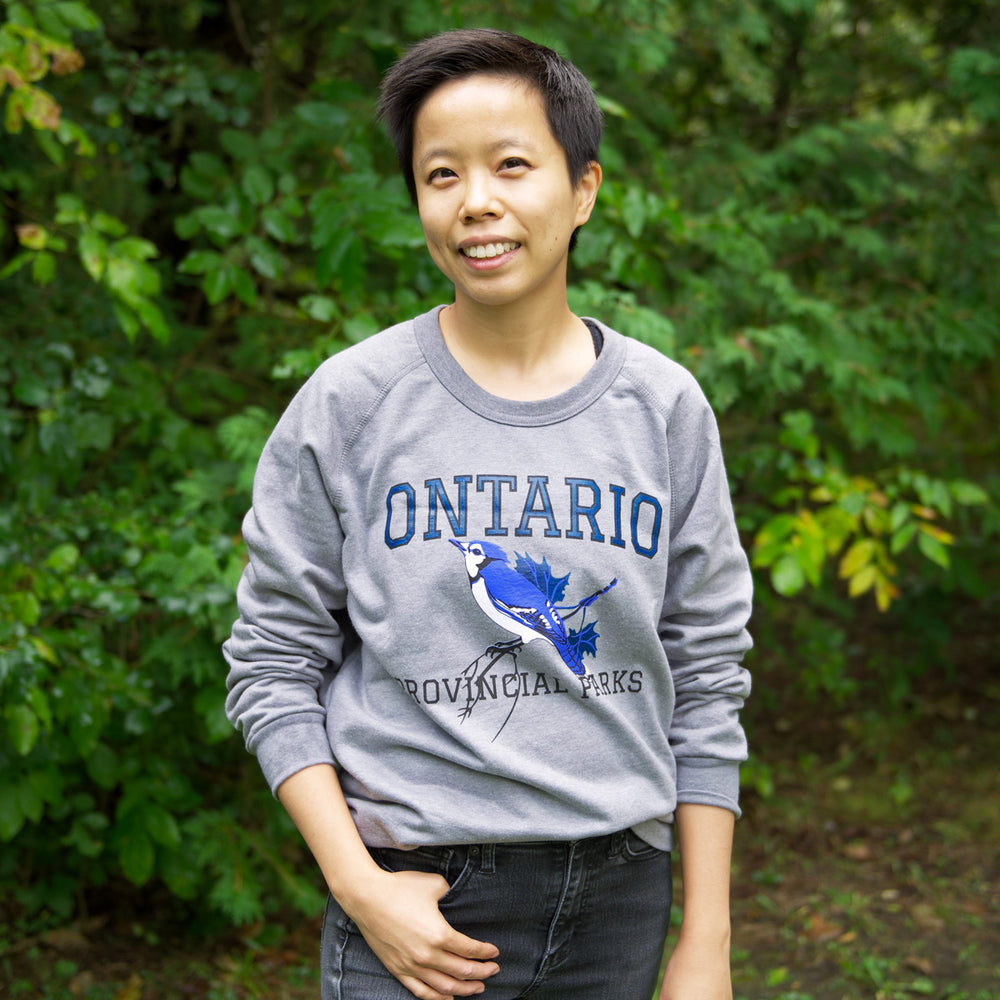 Light Grey Adult vintage crewneck sweatshirt with navy Blue Jay design, worn by adult woman standing in forest. 