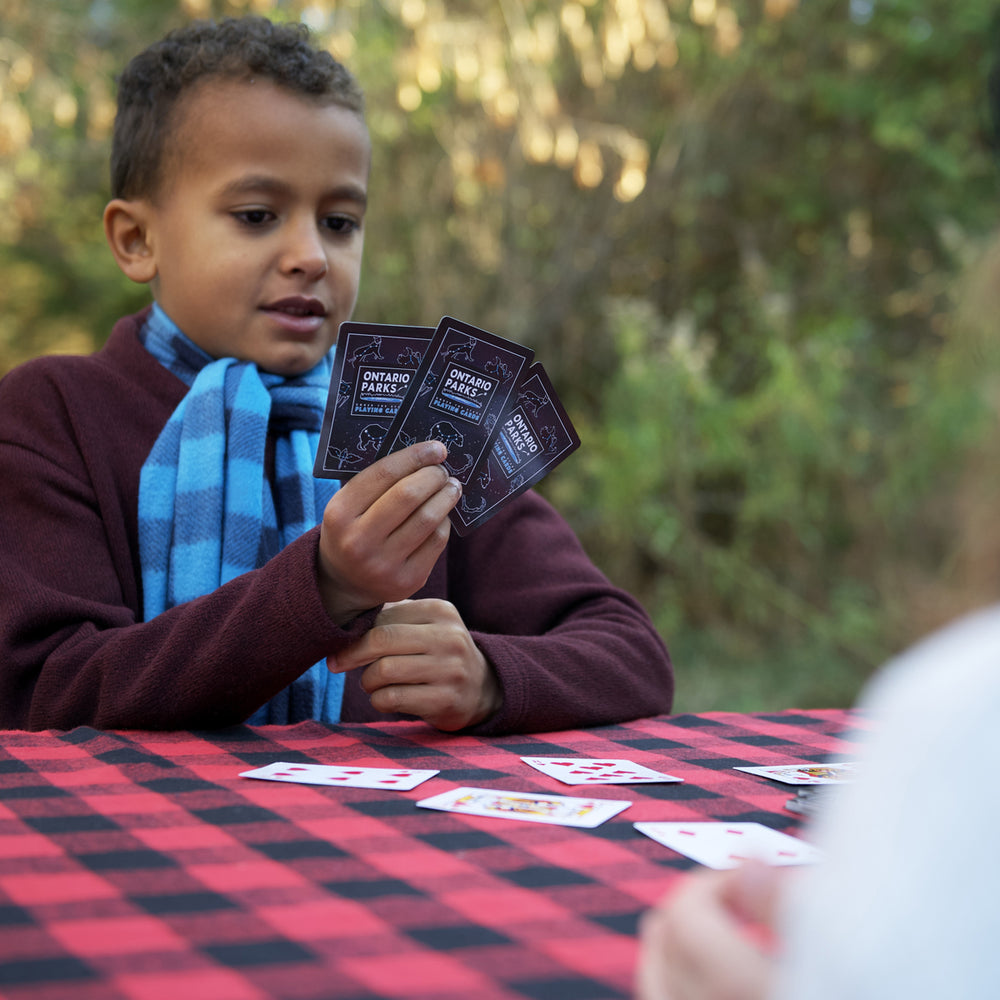 Boy with blue and black scarf sitting at a table playing with Ontario Parks playing cards. Cards are black with Ontario Parks and star constellations on one side. 