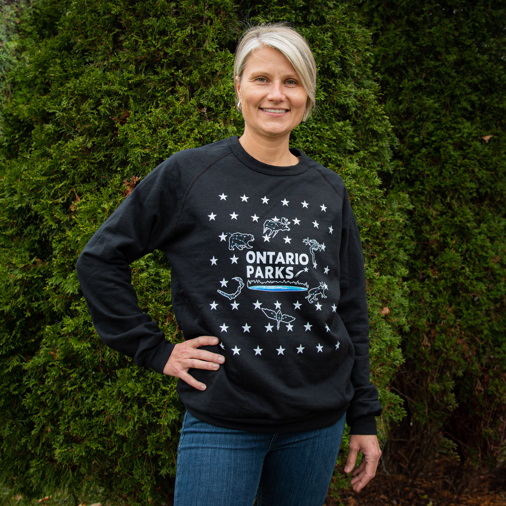 Woman wearing Ontario Parks Black Crew neck featuring star constellations, stars and star patterns in front of evergreen bush