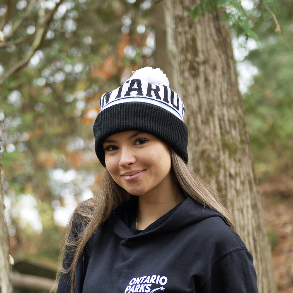 woman standing infront of trees wearing Ontario Parks toque and hoodie. Toque has white fabric with Ontario Parks written with two stars. A black cuff of the hat is folded over itself. 