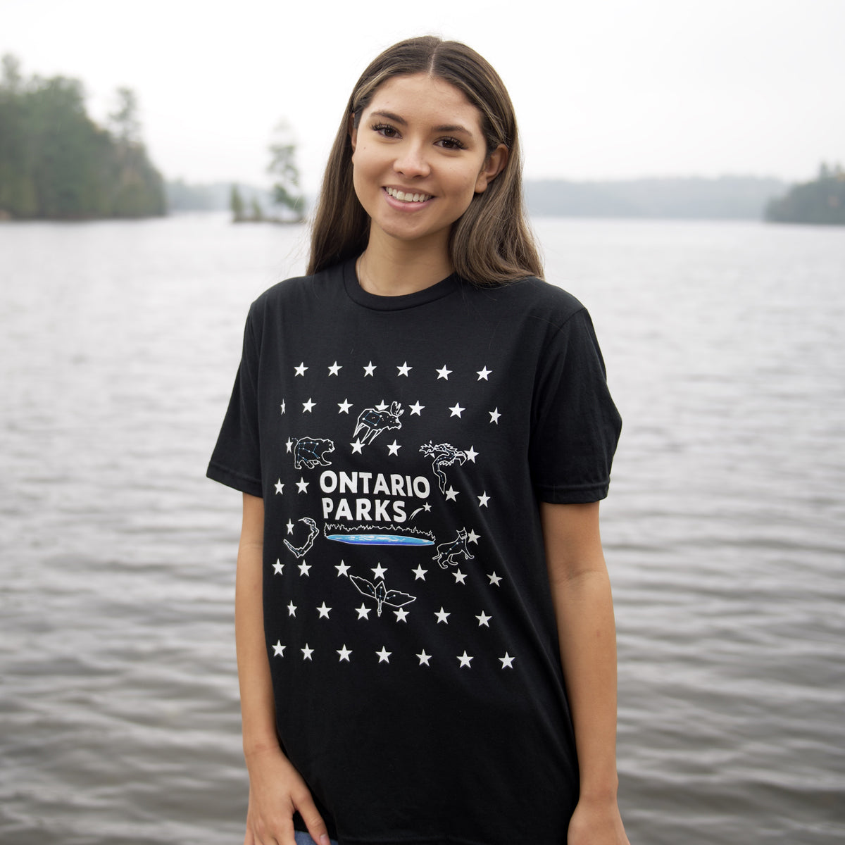 woman standing in front of body of water wearing Ontario Parks black t shirt with stars, star constellations, star patterns 