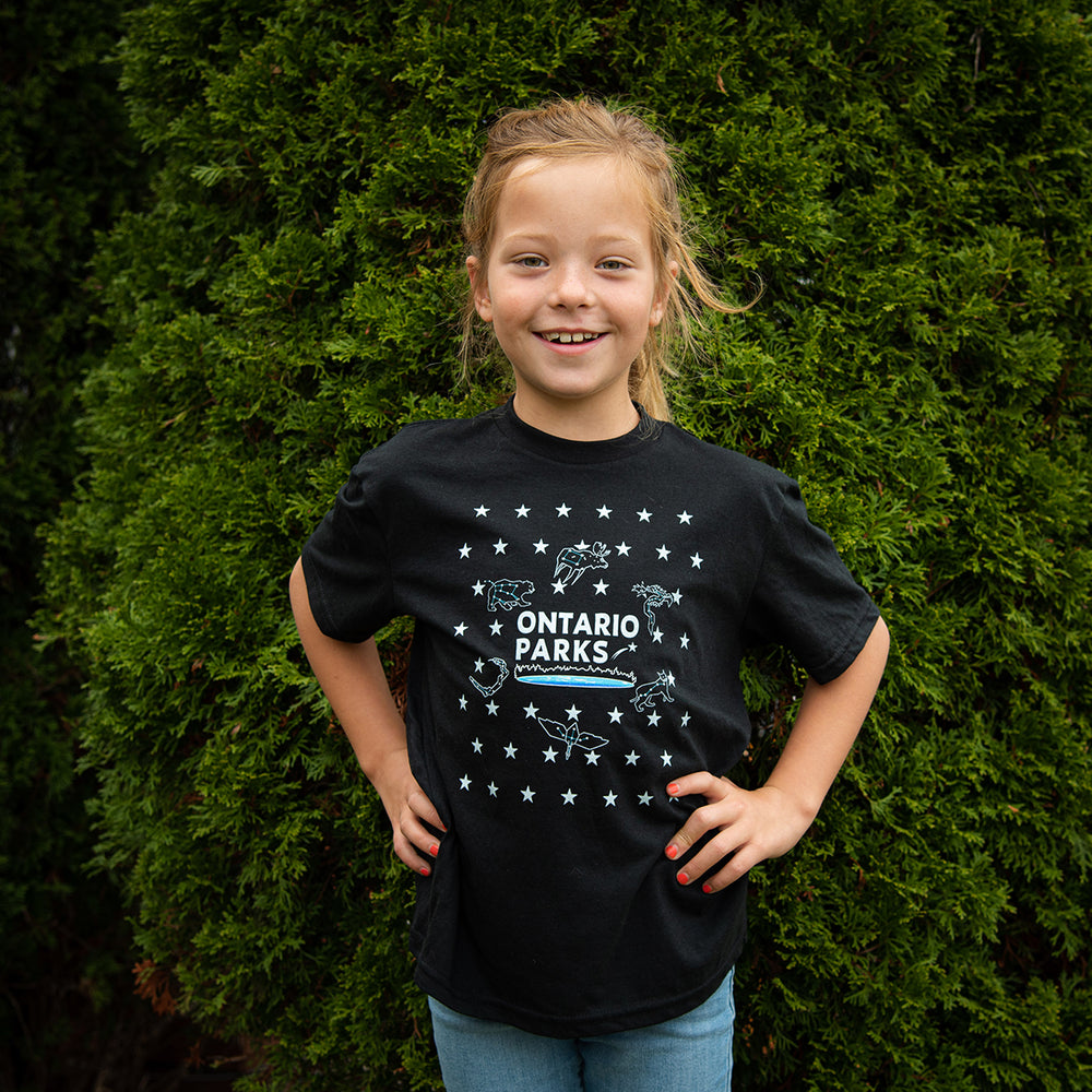 Girl wearing black Ontario Parks t shirt with stars, star constellations and animal constellations in front of evergreen tree. 