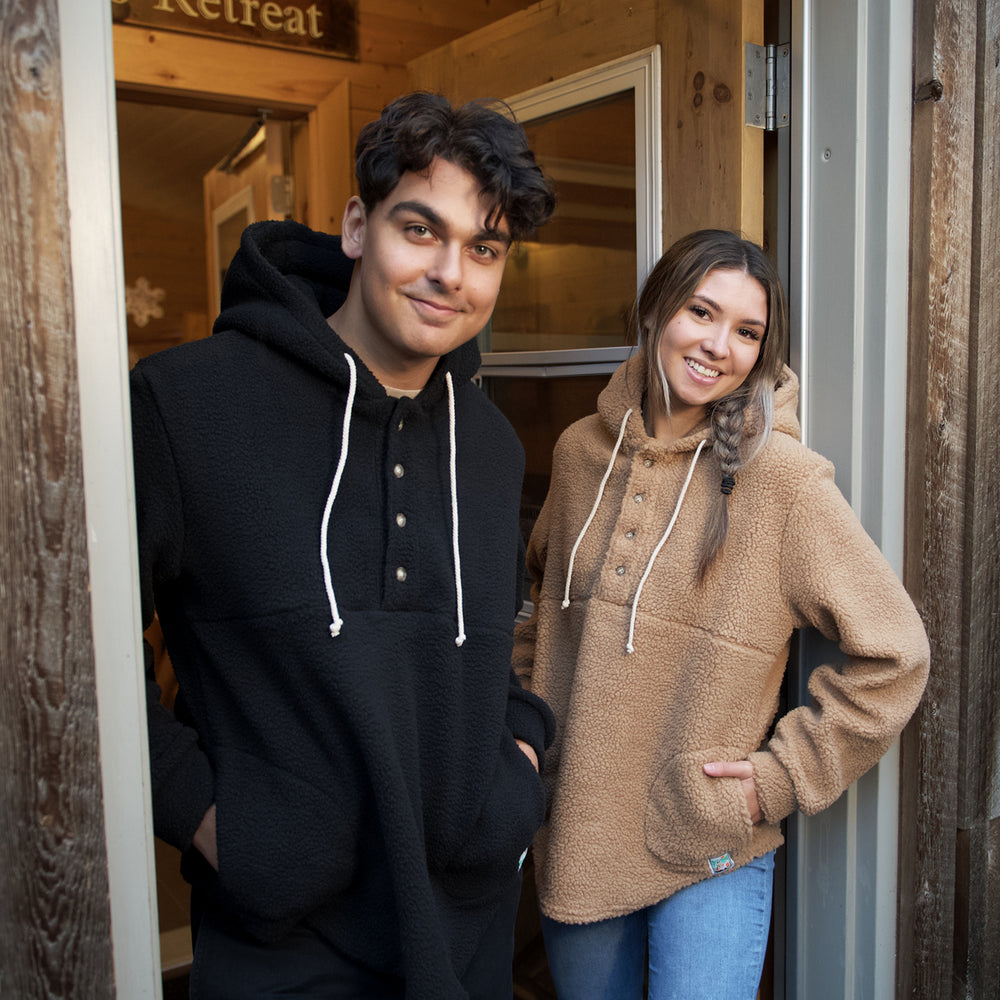 Man and woman standing in doorway of cabin in Muttonhead camp sherpa hoodies. Male is wearing a black sherpa hoodie with white strings around hood and 4 buttons on upper half of sweater with hands in both pockets at sides. Woman is wearing camel sherpa hoodie with white strings and 4 buttons on upper half of sweaters with hands in pockets on both sides.  