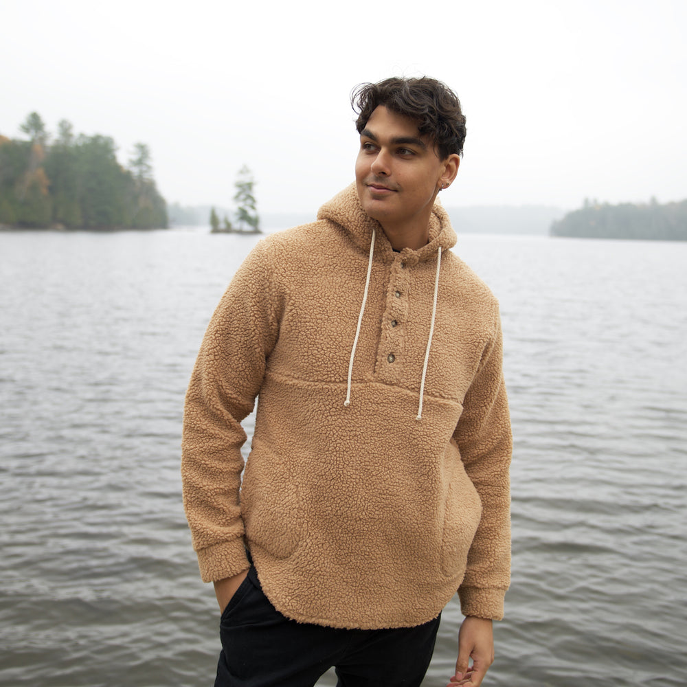 man wearing camel Muttonhead sherpa Hoodie standing in front of body of water. Male has hand in pants pocket. Hoodie has white strings from around hood and 4 buttons around upper half of sweater. 