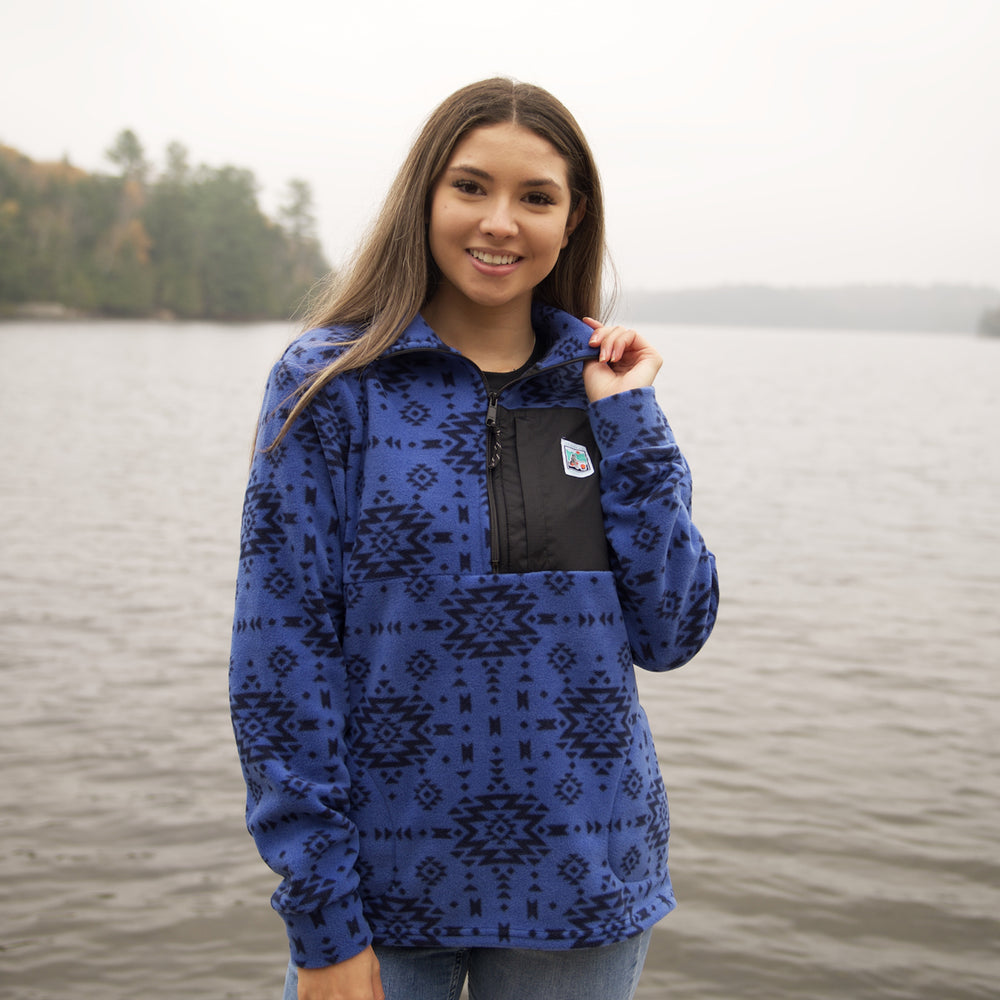 Woman wearing Muttonhead 1/4 zip blue fleece in front of body of water. Woman is touching collar of Sweater. Sweater is blue with black print and a black pocket on top left side with Muttonhead Ontario Parks logo. 