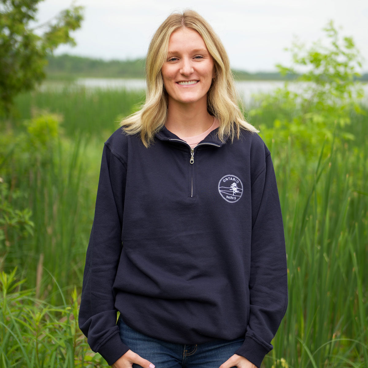 Woman wearing Navy Unisex OP Crest 1/4 Zip Sweatshirt, featuring silver zipper and white embroidered Ontario Parks crest on left chest. 