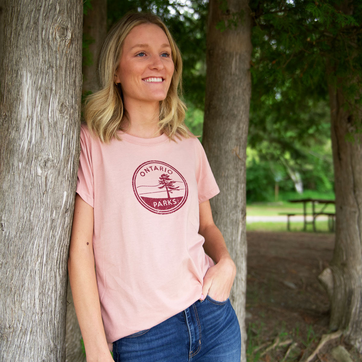 Woman leaning on tree, wearing OP Crest Women's Fit T-shirt (colour Rose). Featuring burgundy Ontario Parks graphic in centre chest.
