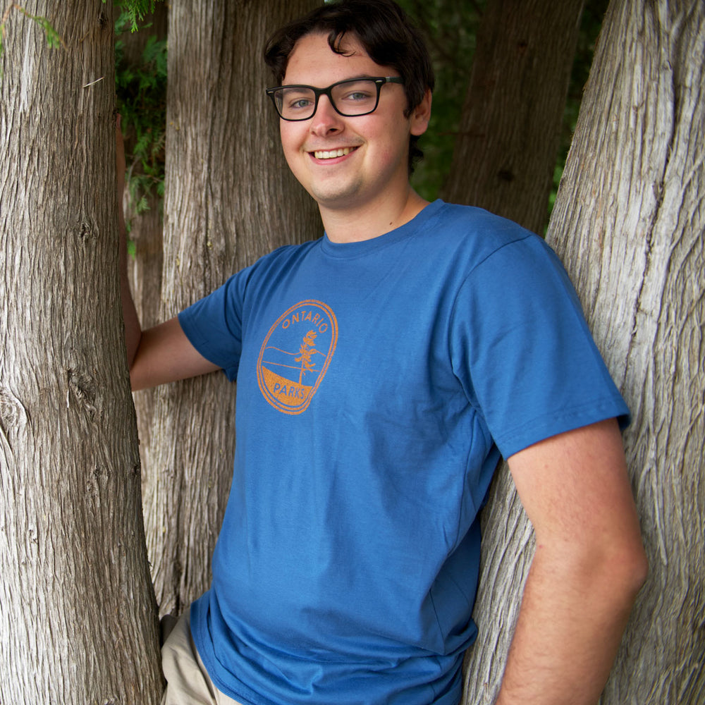 Man leaning on tree, wearing OP Crest Unisex T-shirt in colour Air Force Blue. Featuring orange toned Ontario Parks graphic in centre chest.
