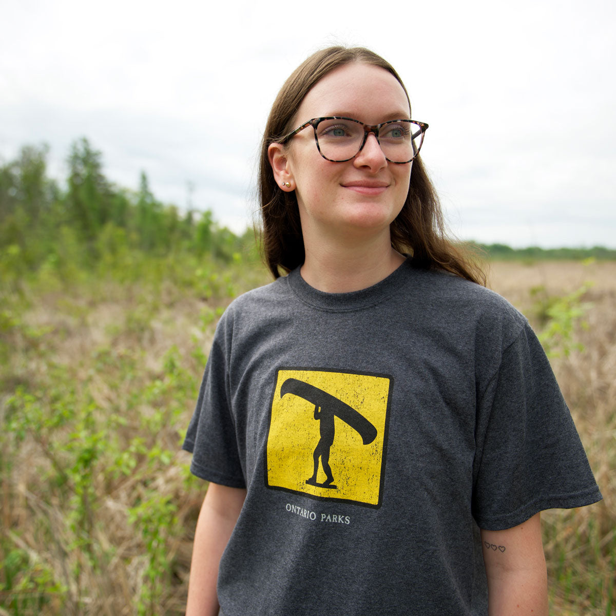 Woman wearing Unisex charcoal grey Park Icon Portage T-shirt, featuring a yellow and black portage logo in a black square on centre chest. 
