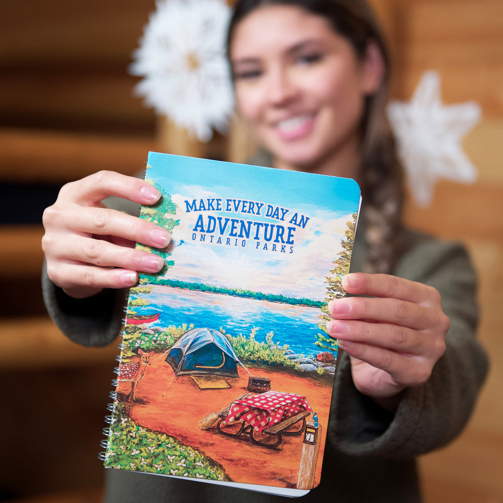 Woman in cozy cabin holding out Ontario Parks Adventure Planner, ring-bound, with hard cover. Cover image shows very colourful camping scene with the text "Make Every Day An Adventure, Ontario Parks".