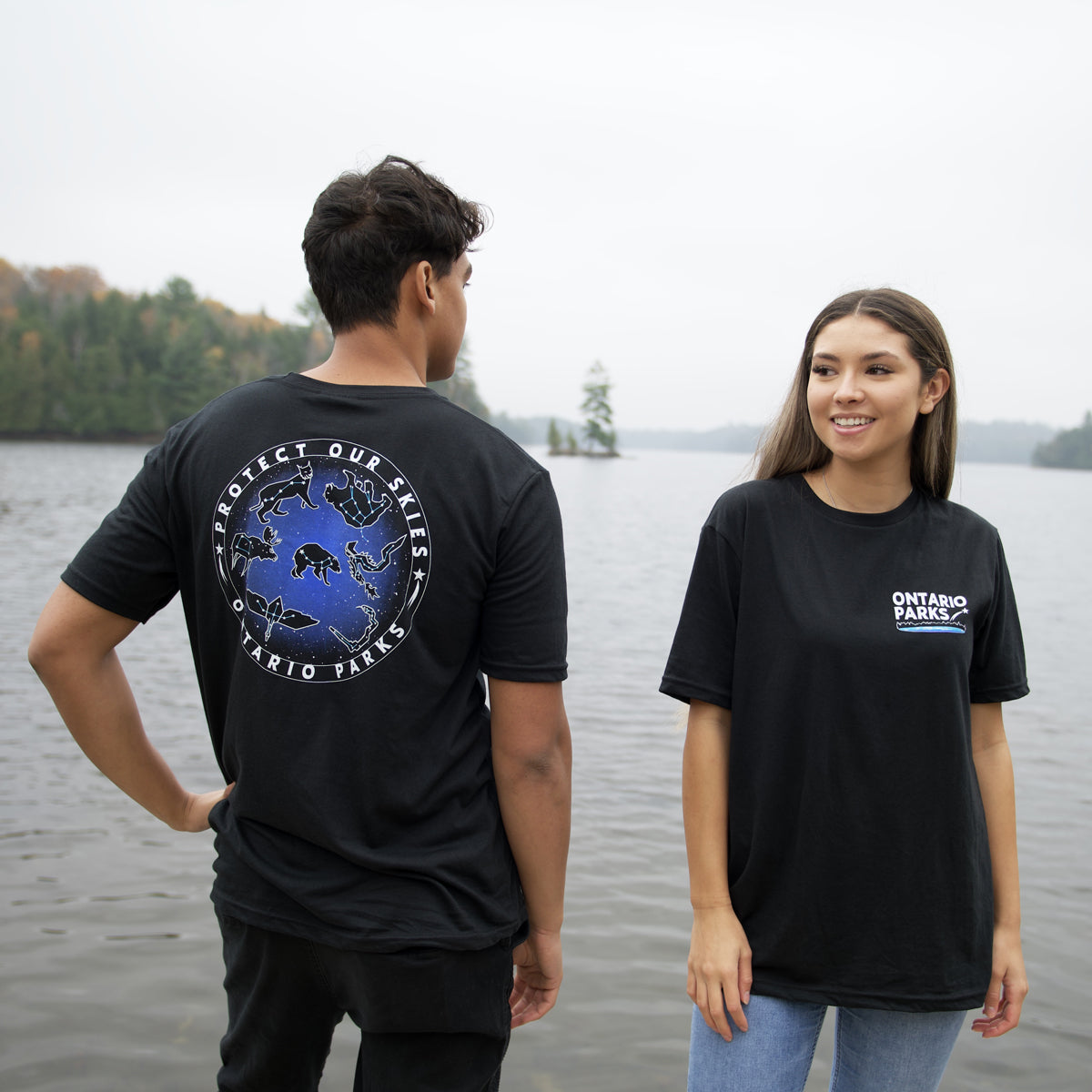 Man standing in front of body of water looking over right shoulder. Wearing Ontario Parks Protect our skies T-Shirt with back to screen showing back of t shirt. Ontario Parks Protect Our Skies text is white and circles around star and animals constellations. Woman is on right side wearing Ontario Parks Protect Our Skies T-shirt facing front. T shirt has Ontario Parks in white text on upper right side with shooting star and blue underline. 