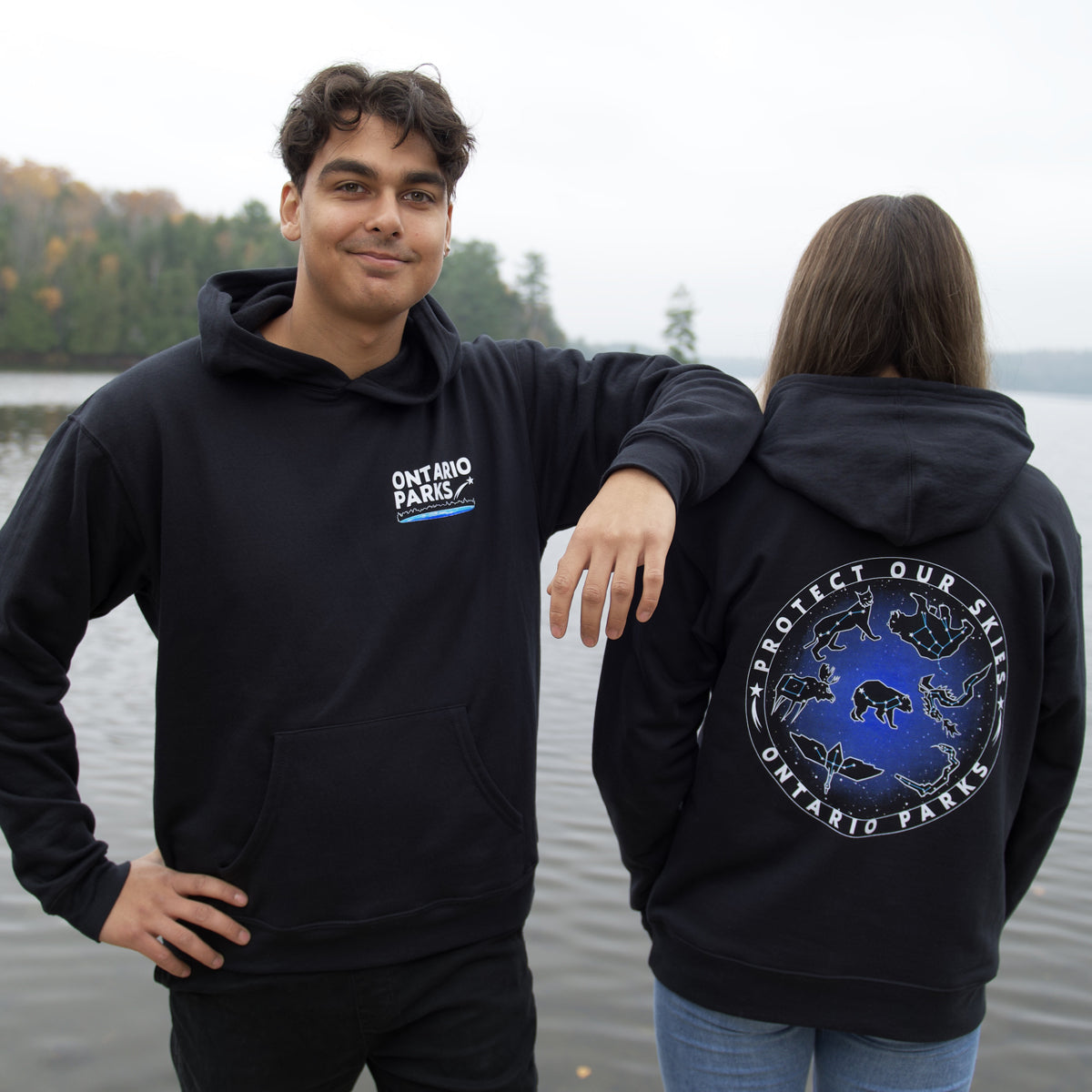 Man and woman standing in front of body of water. Man is on left side wearing Ontario Parks Protect Our Skies hoodie facing front. Hoodie has Ontario Parks in white text on upper right side with shooting star and blue underline. Woman is facing backwards  showing back of hoodie. Ontario Parks Protect Our Skies text is white and circles around star and animals constellations.