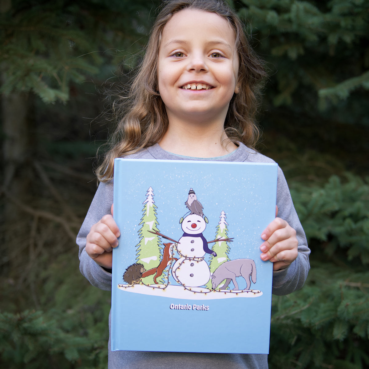 Child wearing grey longsleeve holding Blue Ontario Parks Sketchbook in front of pine trees with animals in winter scene on front cover. Cartoon animals include wolf, porcupine, fox and owl putting light around a snowman.
