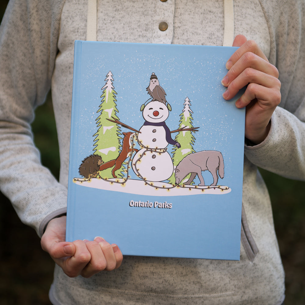 Person wearing light grey sweater holding blue Ontario Parks sketchbook. Blue Ontario Parks Sketchbook features animals in winter scene on front cover. Cartoon animals include wolf, porcupine, fox and owl putting light around a snowman.
