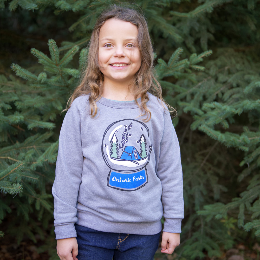 Girl standing in front of pine trees wearing grey Ontario Parks crewneck. Crewneck features winter scene with trees and hot tent in snow globe with Ontario Parks text below.