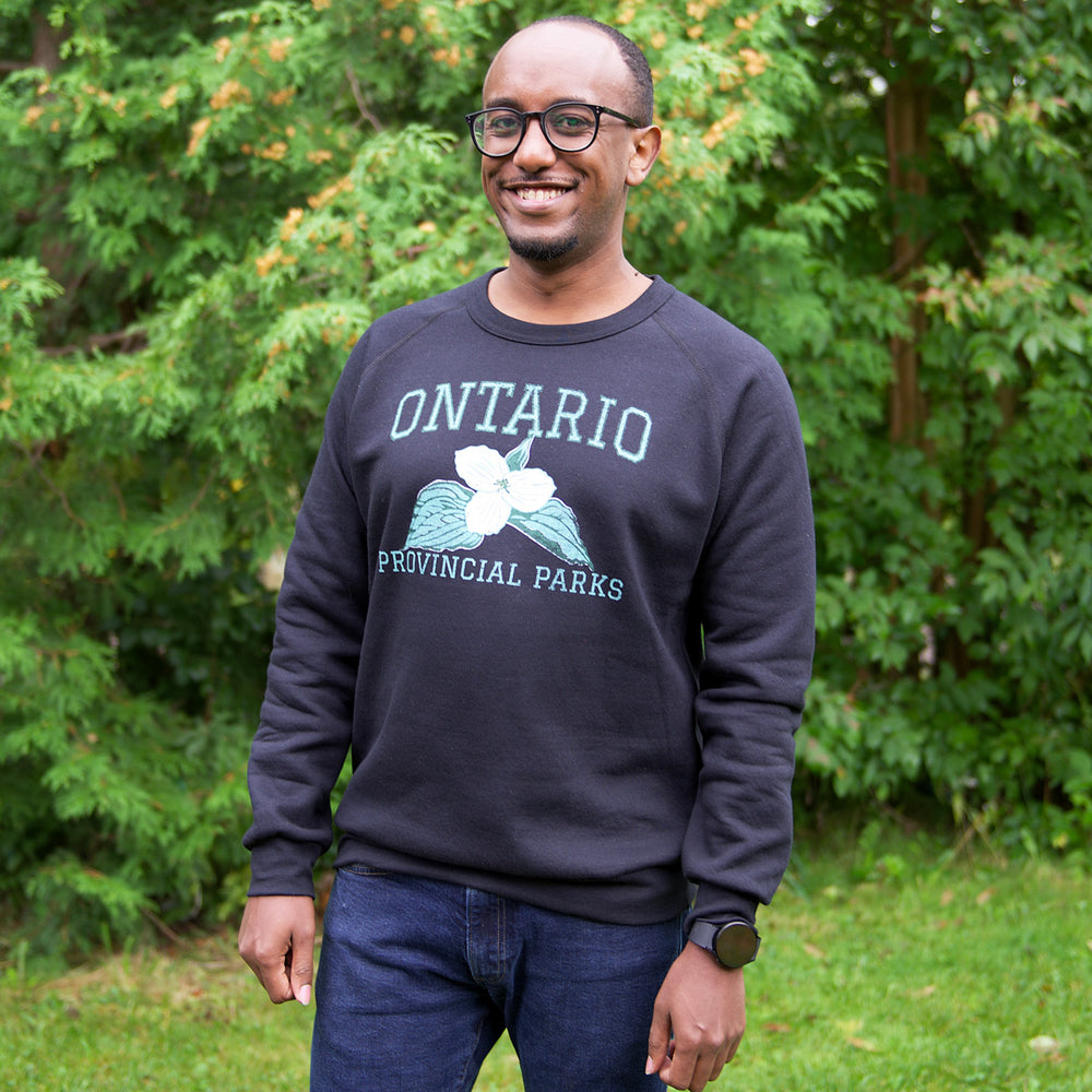 Black Adult vintage crewneck sweatshirt with teal and white Trillium design on centre chest, worn by man standing among trees. 