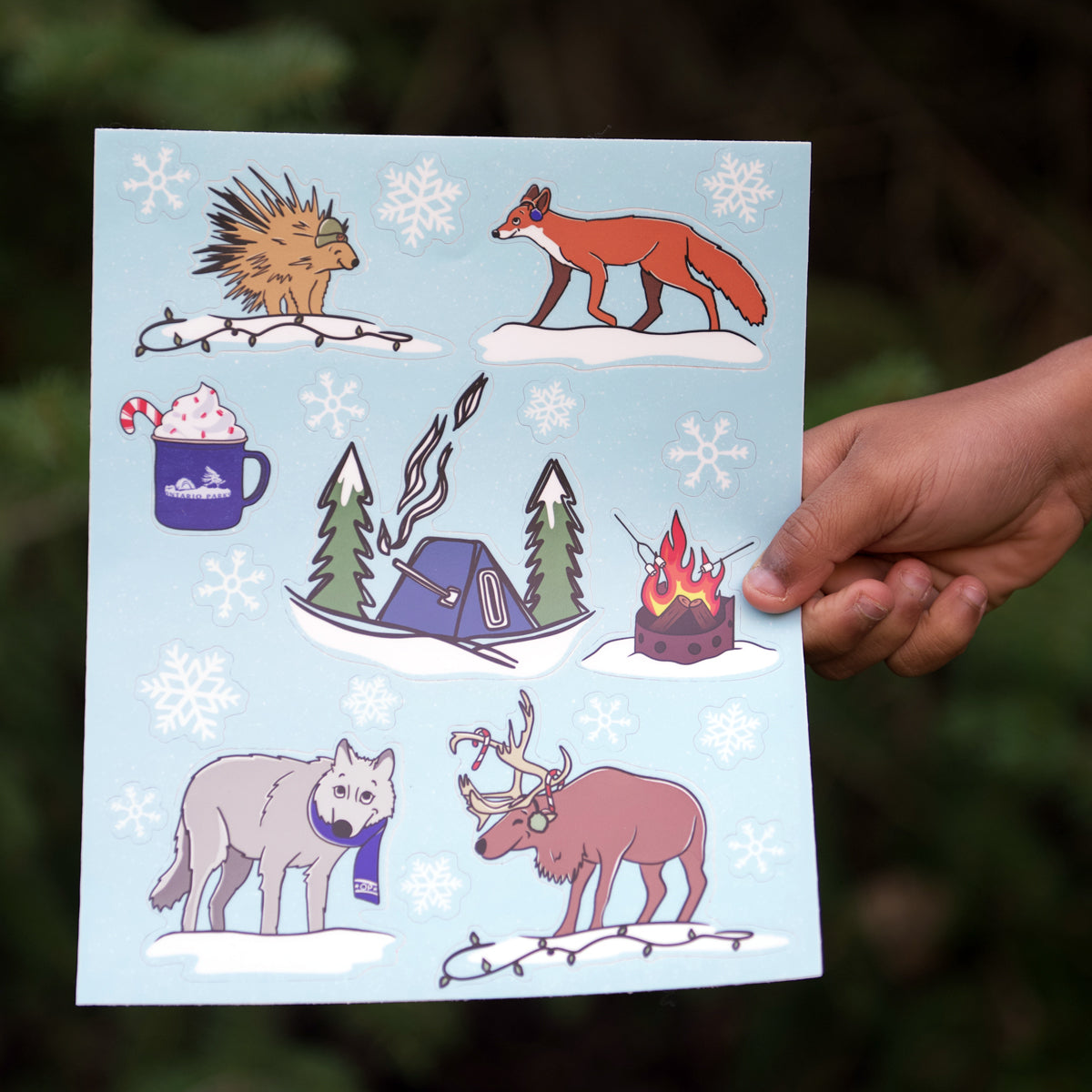 Hand holding out Ontario Parks sticker sheet. Sticker sheet features a porcupine, fox, mug with candy cane, hot tent, campfire with marshmallows, moose, wolf and snowflakes. 