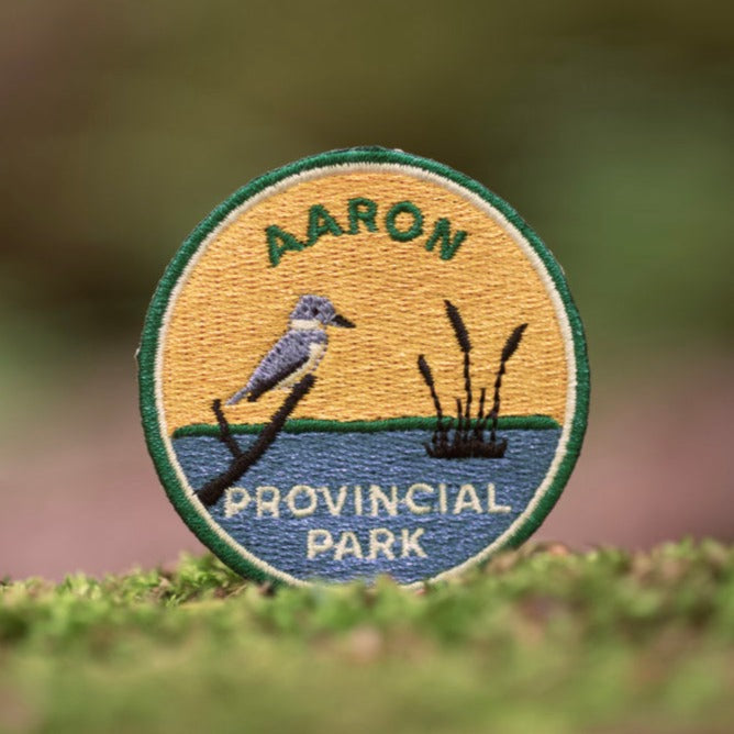 Round embroidered patch with dark green border, of grey bird on branch overlooking blue lake with tall grasses and yellow sky. Text reads Aaron Provincial Park.